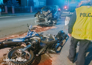 Zdjęcie przedstawiające policjantkę stojącą tyłem. W tle widać rozbity samochód, a obok policjantki rozbity motocykl.