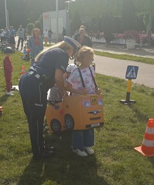 Zdjęcie przedstawiające policjantkę, dwójkę dzieci w tzw. autochodziku. W tle widać osoby uczestniczące w festynie.