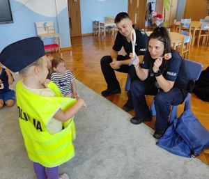 Zdjęcie przedstawiające policjantkę z tarczą drogową, policjanta oraz dziecko w czapce policyjnej oraz kamizelce odblaskowej