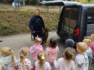 Zdjęcie przedstawiające policjanta oraz dzieci na tle radiowozu. Policjant przedstawia dzieciom psa służbowego i opowiada o jego zadaniach