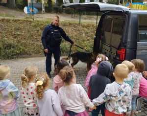 Zdjęcie przedstawiające policjanta, psa służbowego oraz dzieci na tle radiowozu