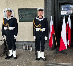 Zdjęcie przedstawiające marynarzy stojących przy tablicy pamiątkowej upamiętniającej ludzi wysiedlonych z Gdyni