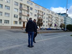 Zdjęcie przedstawiające mężczyznę, który idzie chodnikiem z opaską na oczach i pomaga sobie laską dla niewidomych. Na zdjęciu jest też drugi mężczyzna, stojący tyłem.