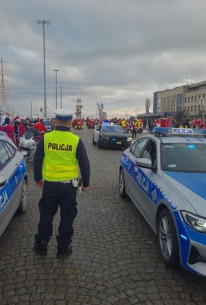 Zdjęcie przedstawiające stojącego tyłem policjanta między dwoma oznakowanymi radiowozami. W tle widać osoby w strojach Mikołajów