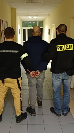 Zdjęcie przedstawiające dwóch policjantów, prowadzących zatrzymanego mężczyznę