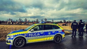 Zdjęcie poglądowe przedstawiające radiowóz policyjny i dwóch policjantów na tle sześciu traktorów