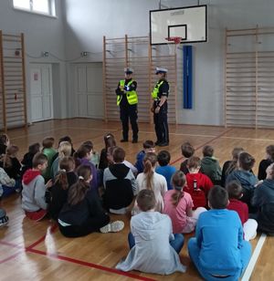 Zdjęcie przedstawiające dwie policjantki, które na sali gimnastycznej rozmawiają z dziećmi na temat bezpieczeństwa w ruchu drogowym. Policjantki stoją przodem do zdjęcia, dzieci siedzą tyłem do zdjęcia.