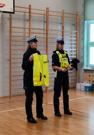 Zdjęcie przedstawiające dwie policjantki, które na sali gimnastycznej promują noszenie elementów odblaskowych, prezentując kamizelki odblaskowe