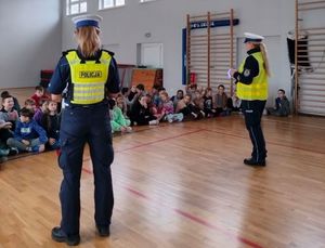Zdjęcie przedstawiające dwie policjantki, które na sali gimnastycznej rozmawiają z dziećmi na temat bezpieczeństwa w ruchu drogowym. Policjantki stoją tyłem do zdjęcia, dzieci siedzą przodem do zdjęcia.