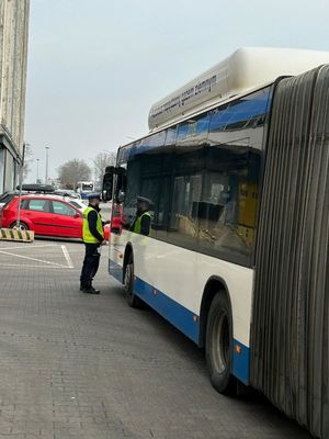 Zdjęcie przedstawiające policjanta podczas kontroli autobusu miejskiego. Na zdjęciu widać autobus i w tle samochód osobowy.