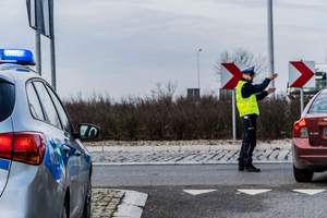 Zdjęcie przedstawiające policjanta ruchu drogowego, który stoi na rondzie i kieruje ruchem kołowym pojazdów. Na zdjęciu widać oznakowany radiowóz policyjny oraz osobowy samochód.