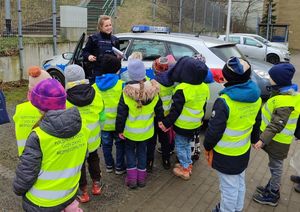 Zdjęcie przedstawiające policjantkę na tle radiowozu oraz dzieci zwrócone tyłem do fotografii. Dzieci stoją na przeciwko policjantki i radiowozu, mają na sobie kamizelki odblaskowe.