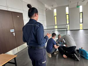 Zdjęcie przedstawiające policjantkę podczas oceny wykonania ćwiczenia przez uczniów. W tle widać pozorantkę siedzącą na podłodze i dwóch uczniów, których zadaniem jest wykonanie poprawnego udzielenia pomocy medycznej.