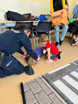 Zdjęcie przedstawiające policjantkę, która pokazuje dzieciom, jak wygląda pozycja bezpieczna podczas udzielania pierwszej pomocy medycznej. Jedno dziecko leży, a drugie pomaga przy ćwiczeniu.