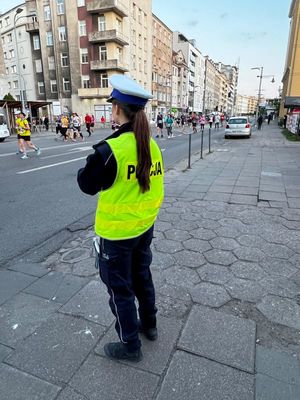 Zdjęcie przedstawiające policjantkę ruchu drogowego, która zabezpiecza bieg. W tle widać uczestników biegu.