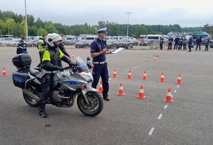 Zdjęcie przedstawiające policjanta na motocyklu, policjanta egzaminującego konkurencję oraz w tle innych policjantów i pojazy