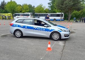 Zdjęcie przedstawiające policjanta w radiowozie, policjanta egzaminującego, a w tle osoby obserwujące konkurencję, wśród nich policjantów. Ponadto widać zaparkowane pojazdy.