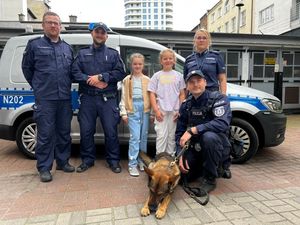 Zdjęcie przedstawiające policjantkę, trzech policjantów, dwie dziewczynki oraz psa służbowego. Wszyscy stoją na tle radiowozu policyjnego.