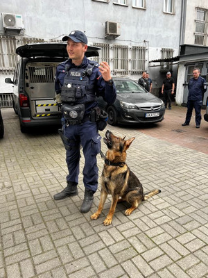 Zdjęcie przedstawiające policjanta z psem służbowym na tle radiowozu policyjnego.