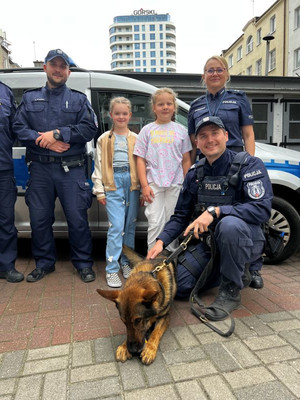 Zdjęcie przedstawiające policjantkę, dwóch policjantów, dwie dziewczynki oraz psa służbowego. Wszyscy stoją na tle radiowozu policyjnego.