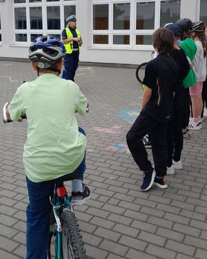 Zdjęcie przedstawiające policjantkę ruchu drogowego, która na placu szkolnym przypomina dzieciom o bezpieczeństwie w ruchu drogowym. Na zdjęciu widać grupę stojących uczniów oraz jednego ucznia na rowerze.