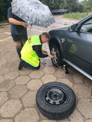 Zdjęcie przedstawiające policjanta, który wymienia koło w samochodzie. Na zdjęciu widać mężczyznę, który stoi przy policjancie i trzyma parasol.