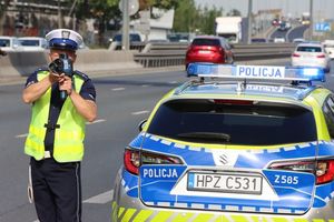 Zdjęcie przedstawiające policjanta drogówki podczas pomiaru prędkości. Policjant stoi przy oznakowanym radiowozie.