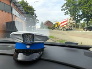 Zdjęcie przedstawiające czapkę policjanta na desce rozdzielczej radiowozu. W tle widać pojazdy i pieszych przed zamkniętym szlabanem.