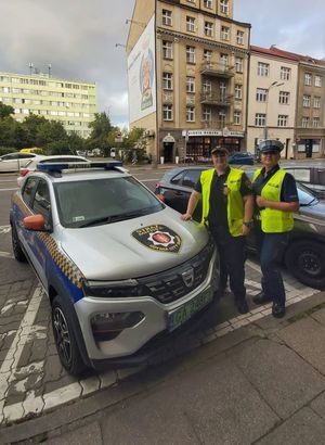 Zdjęcie przedstawiające policjantkę i strażniczkę miejską na tle samochodu Straży Miejskiej.