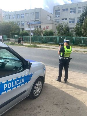 Zdjęcie przedstawiające policjanta, który wykonuje pomiar prędkości samochodom. W tle widać szkołę oraz uczniów. Na zdjęciu widać również radiowóz policyjny.