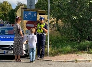 Zdjęcie przedstawiające policjantkę, która rozmawia z dzieckiem. Obok dziecka stoi kobieta. Na zdjęciu widać znaki drogowe oraz radiowóz policyjny.