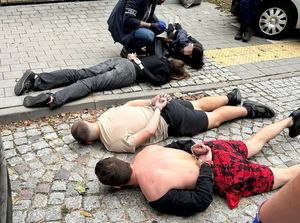 Zdjęcie przedstawiające policjanta kucającego przy czterech osobach, leżących na ziemi.