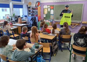 Zdjęcie przedstawiające policjantkę, która rozmawia z uczniami na temat odblasków i bezpieczeństwa na drodze. Policjantka stoi naprzeciw dzieci, trzyma w rękach kamizelkę odblaskową oraz tarczę drogową.