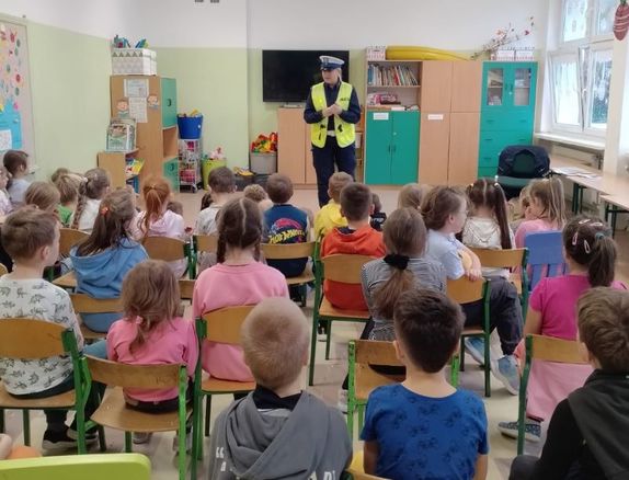 Zdjęcie przedstawiające policjantkę w kamizelce odblaskowej, podczas rozmowy z uczniami. Policjantka stoi przodem do dzieci, dzieci siedzą tyłem do zdjęcia, skierowane na policjantkę.