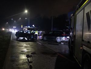 Zdjęcie przedstawiające uszkodzone dwa samochody na jezdni. Z prawej strony widać wóz strażacki, a w tle radiowóz policyjny oraz policjanta.