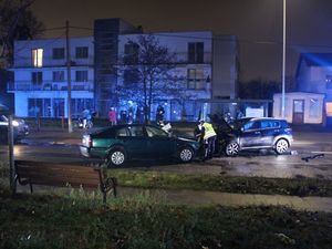 Zdjęcie przedstawiające dwa uszkodzone samochody na jezdni oraz policjanta. W tle widać kilka osób stojących na chodniku.