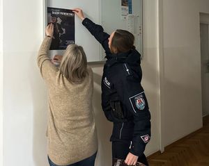 Zdjęcie przedstawiające kobietę oraz dzielnicową, przywieszające plakat, dotyczący naboru do Policji.