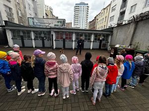 Zdjęcie przedstawiające dzieci, które oglądają pokaz umiejętności psa służbowego. Na zdjęciu widać też przewodnika psa.