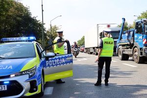 Zdjęcie przedstawiające policjantów drogówki na tle radiowozu na drodze oraz inne pojazdy.