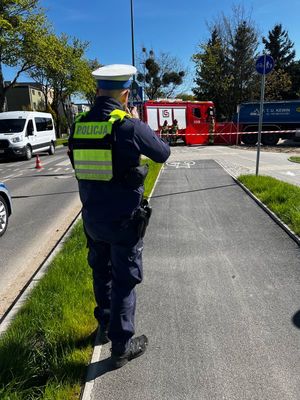 Zdjęcie przedstawiające policjanta ruchu drogowego podczas wykonywania dokumentacji fotograficznej z miejsca wypadku. Na zdjęciu widać również wóz strażacki, pojazd ciężarowy oraz jadący ulicą samochód.