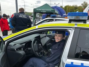 Zdjęcie przedstawiające policjantkę w radiowozie policyjnym. W tle widać stojącego tyłem strażnika miejskiego.