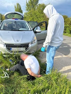 Zdjęcie przedstawiające siedzącego przed pojazdem zatrzymanego mężczyznę. Na zdjęciu widać policjanta, stojącego przy mężczyźnie.