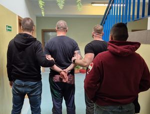 Zdjęcie przedstawiające dwóch policjantów prowadzących zatrzymanego mężczyznę. Za nimi stoi trzeci policjant.