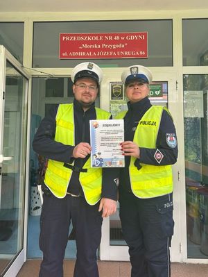 Zdjęcie przedstawiające policjantkę oraz policjanta stojących przed wejściem do placówki przedszkolnej i trzymających w rękach podziękowanie.