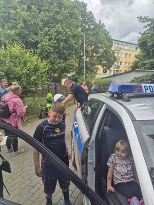Zdjęcie przedstawiające jedno dziecko w radiowozie, drugie stojące przy siedzeniu pasażera. W tle widać policjanta i cztery osoby, które są uczestnikami festynu.