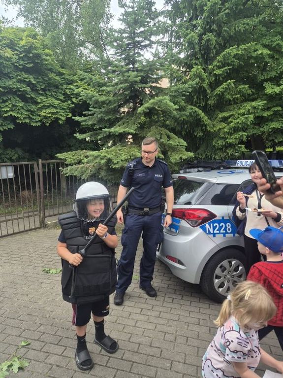 Zdjęcie przedstawiające dzieci przy policjancie na tle radiowozu. Jedno z dzieci ma na sobie wyposażenie policyjne.