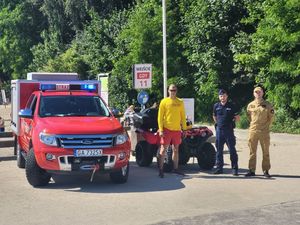 Zdjęcie przedstawiające policjanta, funkcjonariusza Państwowej Straży Pożarnej oraz ratownika WOPR na tle radiowozu straży pożarnej, przy wejściu na plaże miejską nr 11.