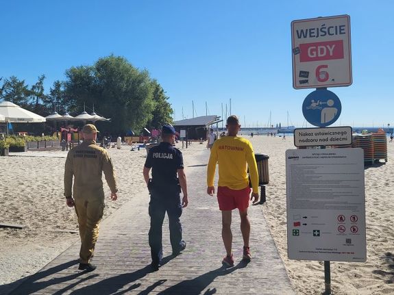Zdjęcie przedstawiające policjanta, funkcjonariusza Państwowej Straży Pożarnej oraz ratownika WOPR patrolujących plaże miejską od wejścia nr 6.