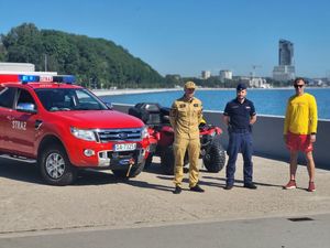 Zdjęcie przedstawiające policjanta, funkcjonariusza Państwowej Straży Pożarnej oraz ratownika WOPR na bulwarze przy plaży miejskiej.