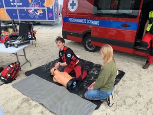 Zdjęcie przedstawiające ratownika prezentującego udzielanie pierwszej pomocy na fantomie podczas festynu na plaży miejskiej. Na zdjęciu widać również młodą kobietę.